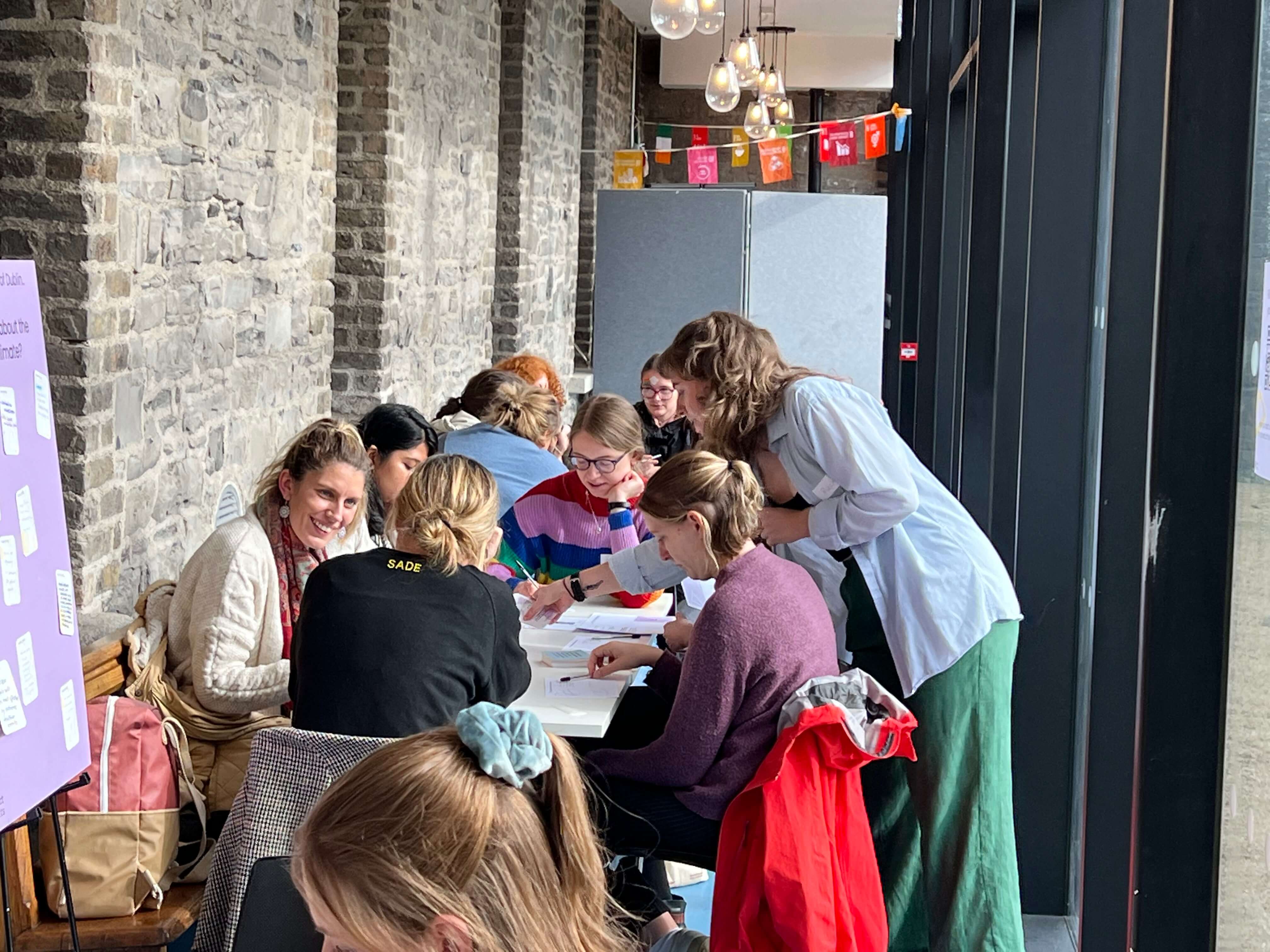 A young woman leans over a table of women to point at a worksheet on the table. The group at the table are completing a worksheet activity. Some are writing or are deep in thought, some are laughing and discussing the activity. A similar group set up can be seen in the background