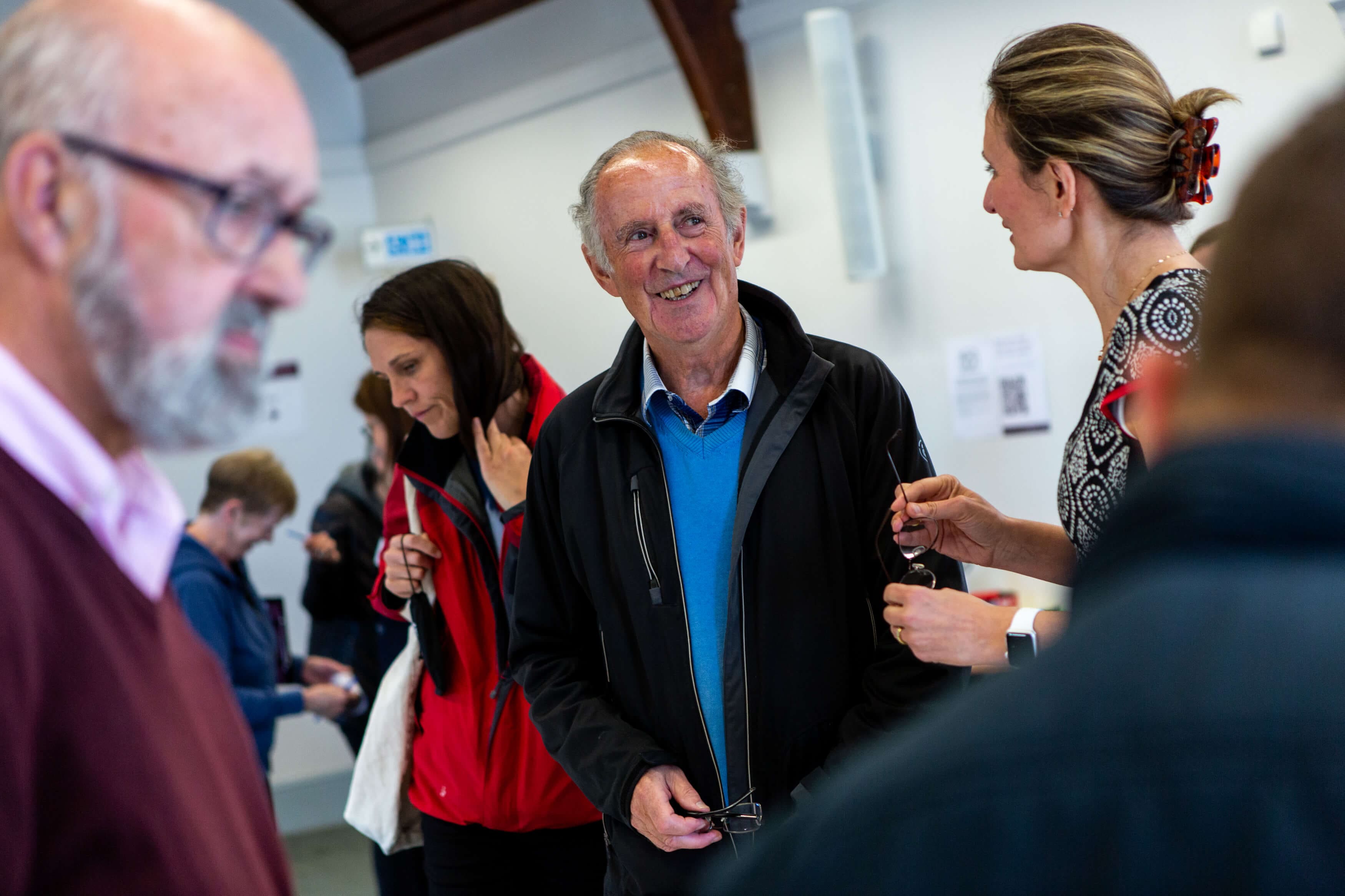An older man and woman are in conversation. The man is smiling and laughing at something the woman has said. Other people in the background are viewing exhibition materials