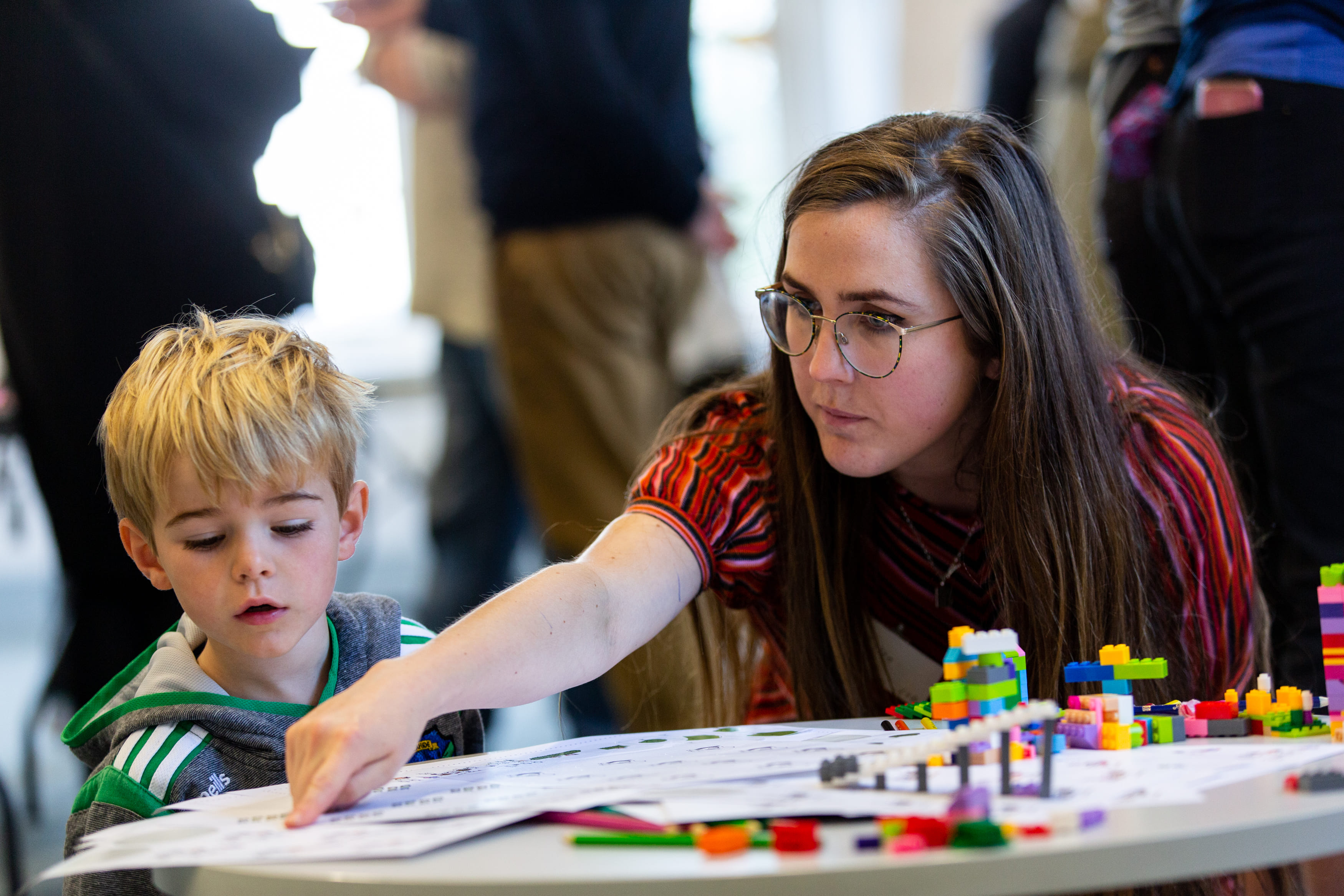 A young boy and a woman sit at a low table. The table has activity worksheets and lego. The woman points at something on the worksheet, explaining it to the child