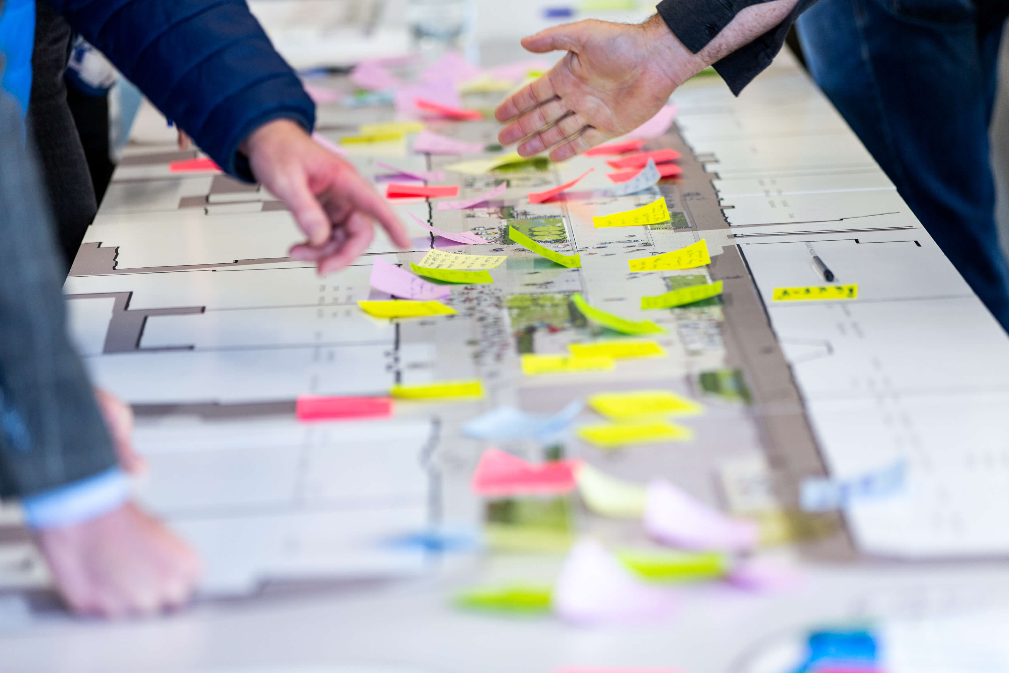 Three pairs of hands gesture and point towards a large map on a table. A street layout with trees and other landscape features can be seen. Sticky notes of various colours cover the length of the map