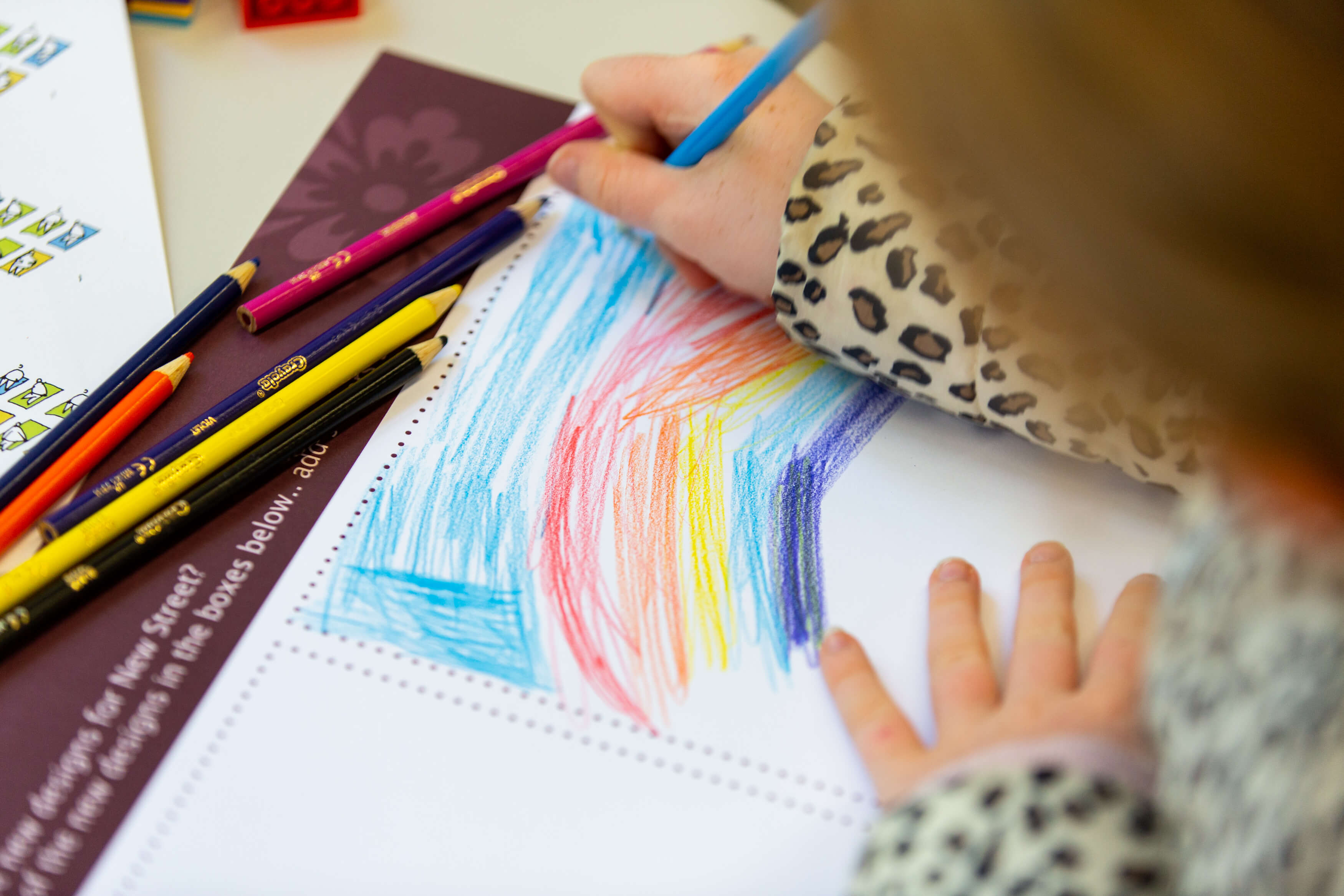 Overhead view of a young girl drawing a rainbow and colouring a blue sky on a workshop activity sheet. Other colouring pencils and stickers sit on the table beside her