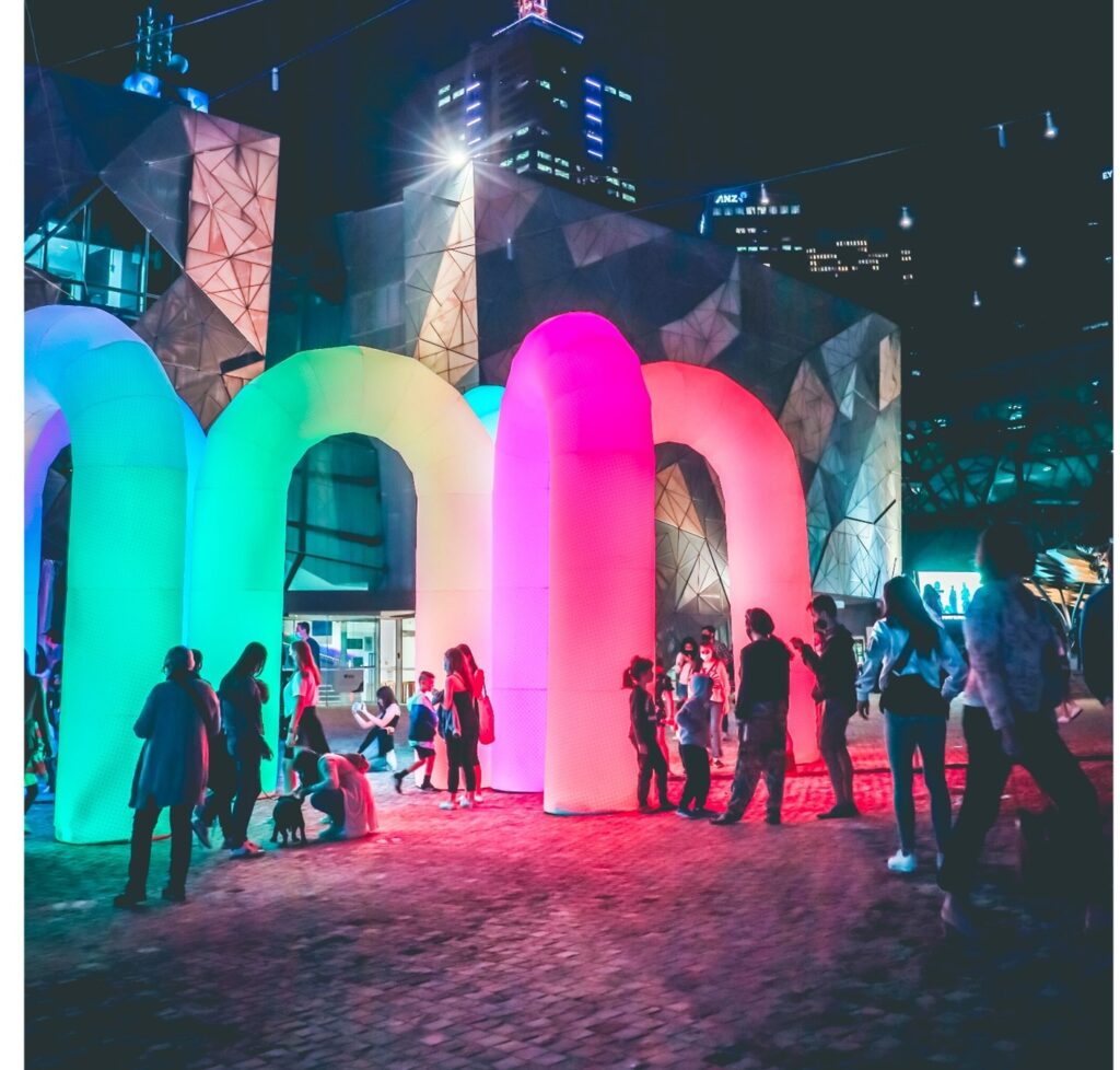 A public art installation at night, large tube like arches lit up in different colours stand in a central square, a crowd gathers around looking at the art and taking pictures. City buildings can be seen in the background