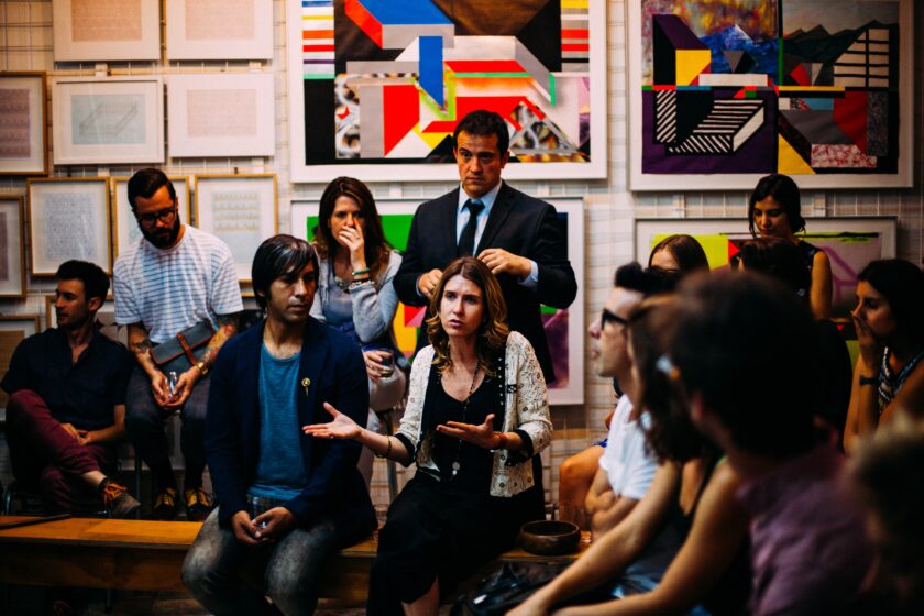 A group of men and women sit or stand in a circle. A woman in the centre of the image is speaking animatedly, the rest listen. They are in a gallery space with a variety of colourful paintings and sketches hung on the walls behind them